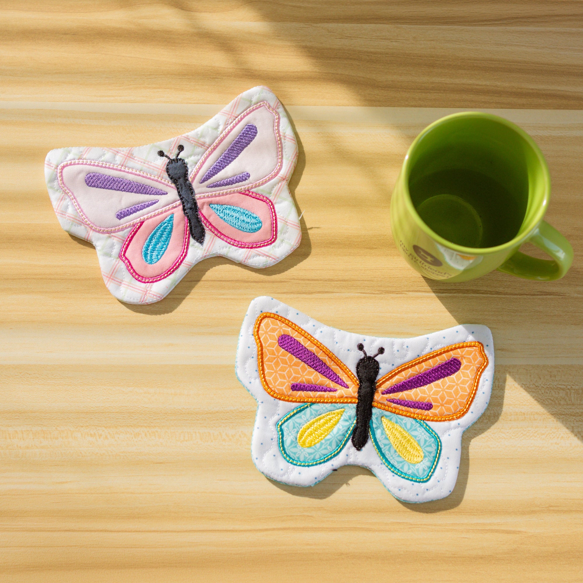 Two colorful butterfly coasters on a wooden surface with a green mug.