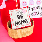 Valentine's Day-themed pillow with 'Be Mine' text in a basket on a red background.