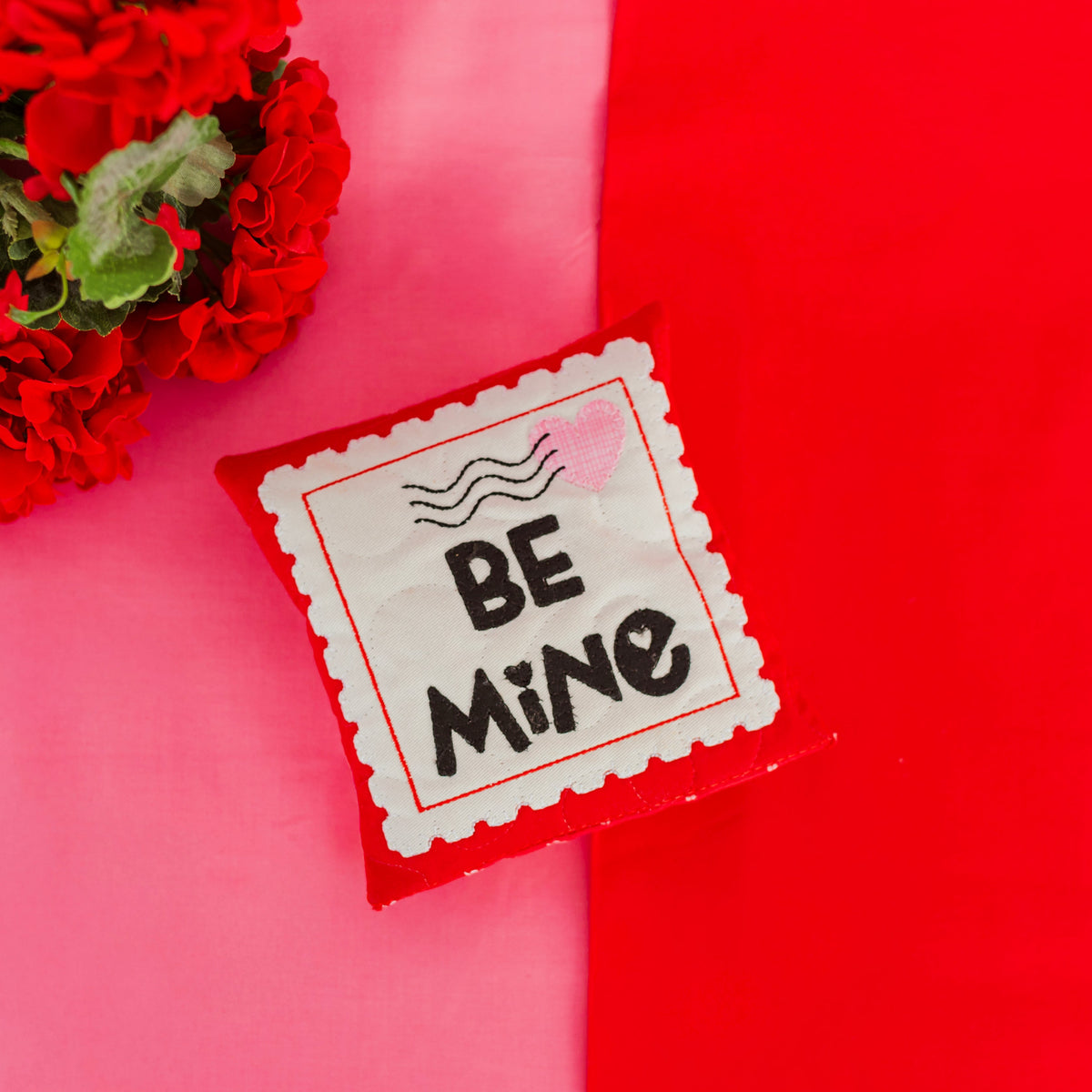 Valentine's Day card with 'Be Mine' text on a pink background with red flowers.
