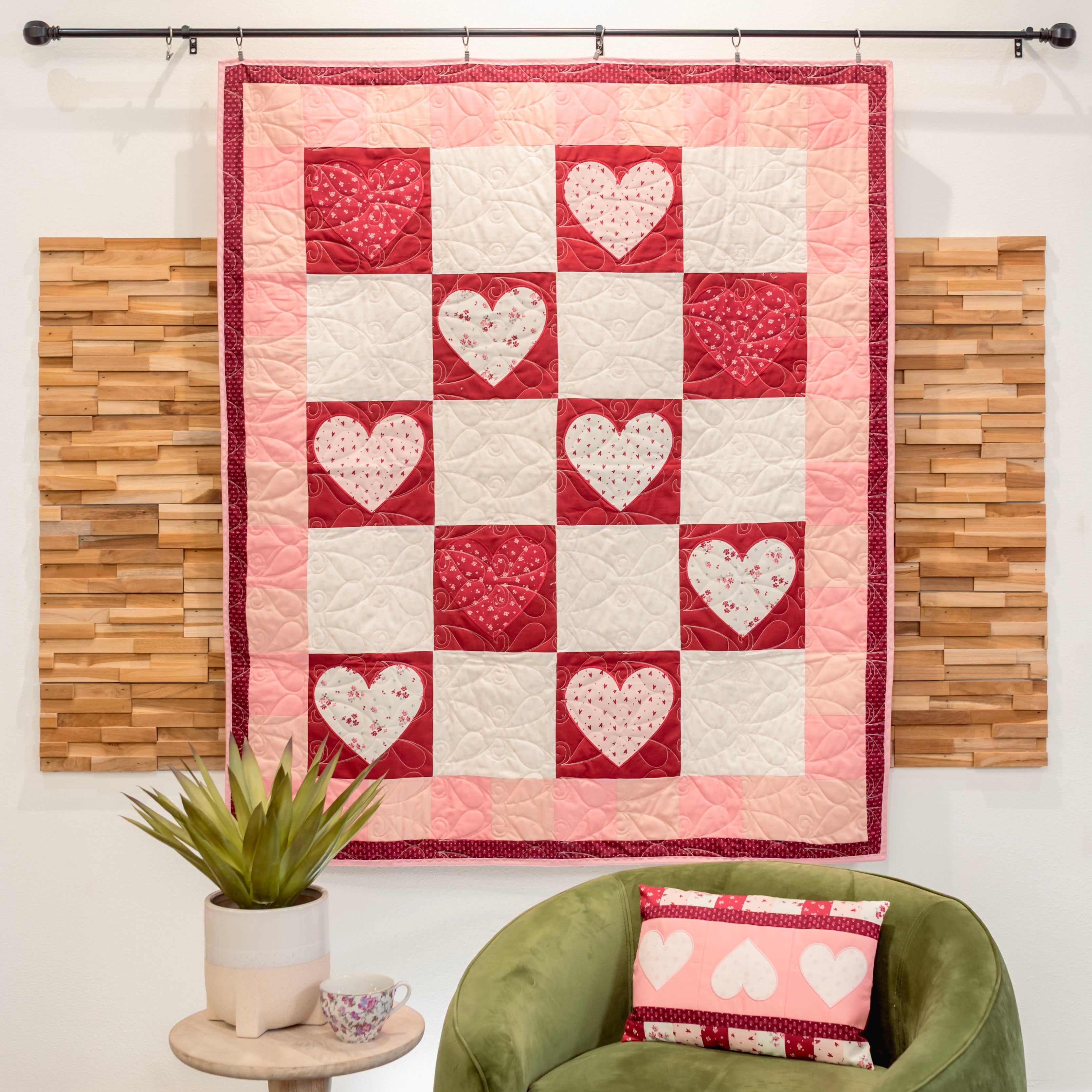 Quilt with heart pattern hanging on a wall above a green chair with a matching pillow.