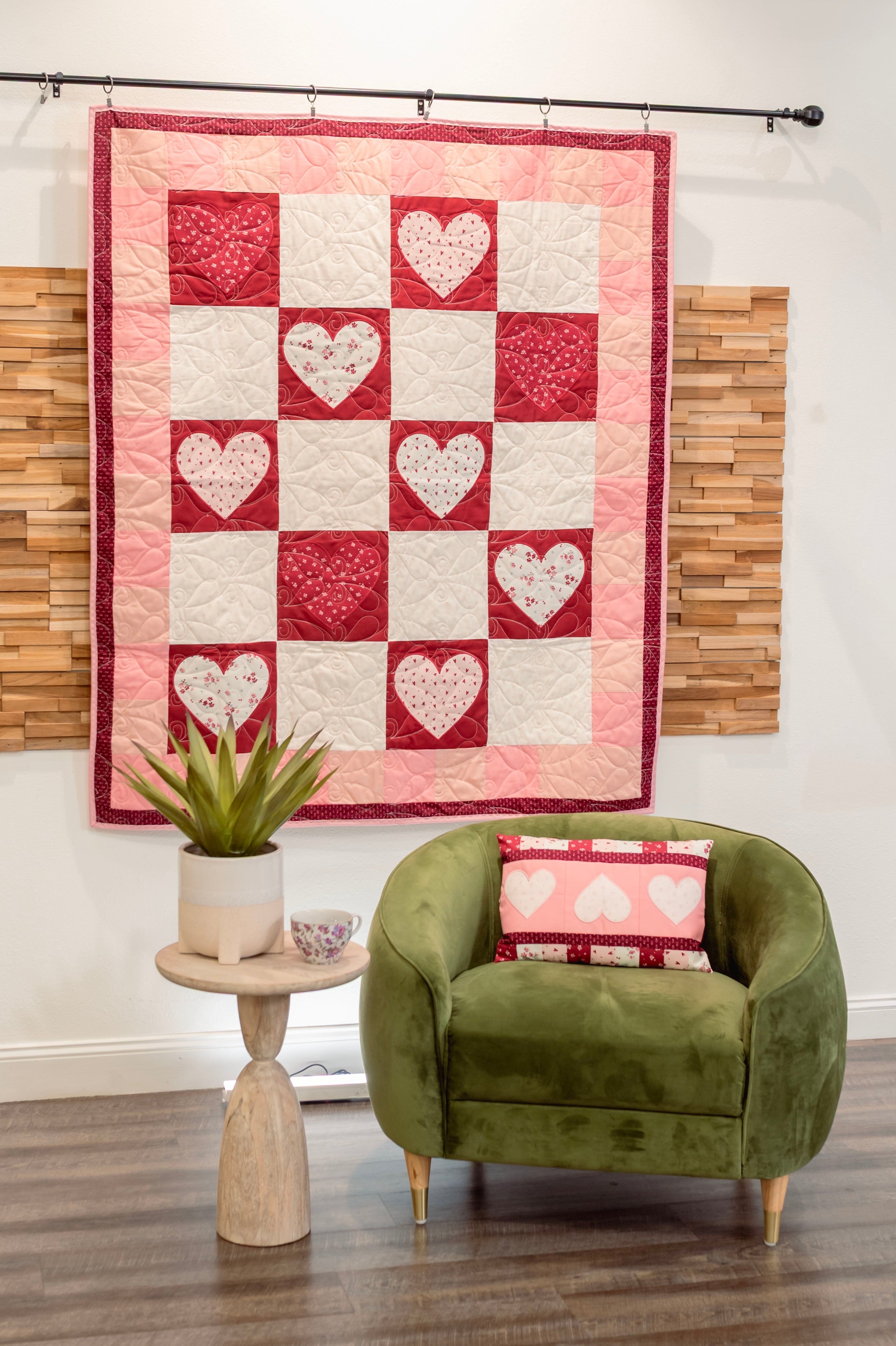 Quilt with heart pattern hanging on a wall above a green armchair with a matching pillow.