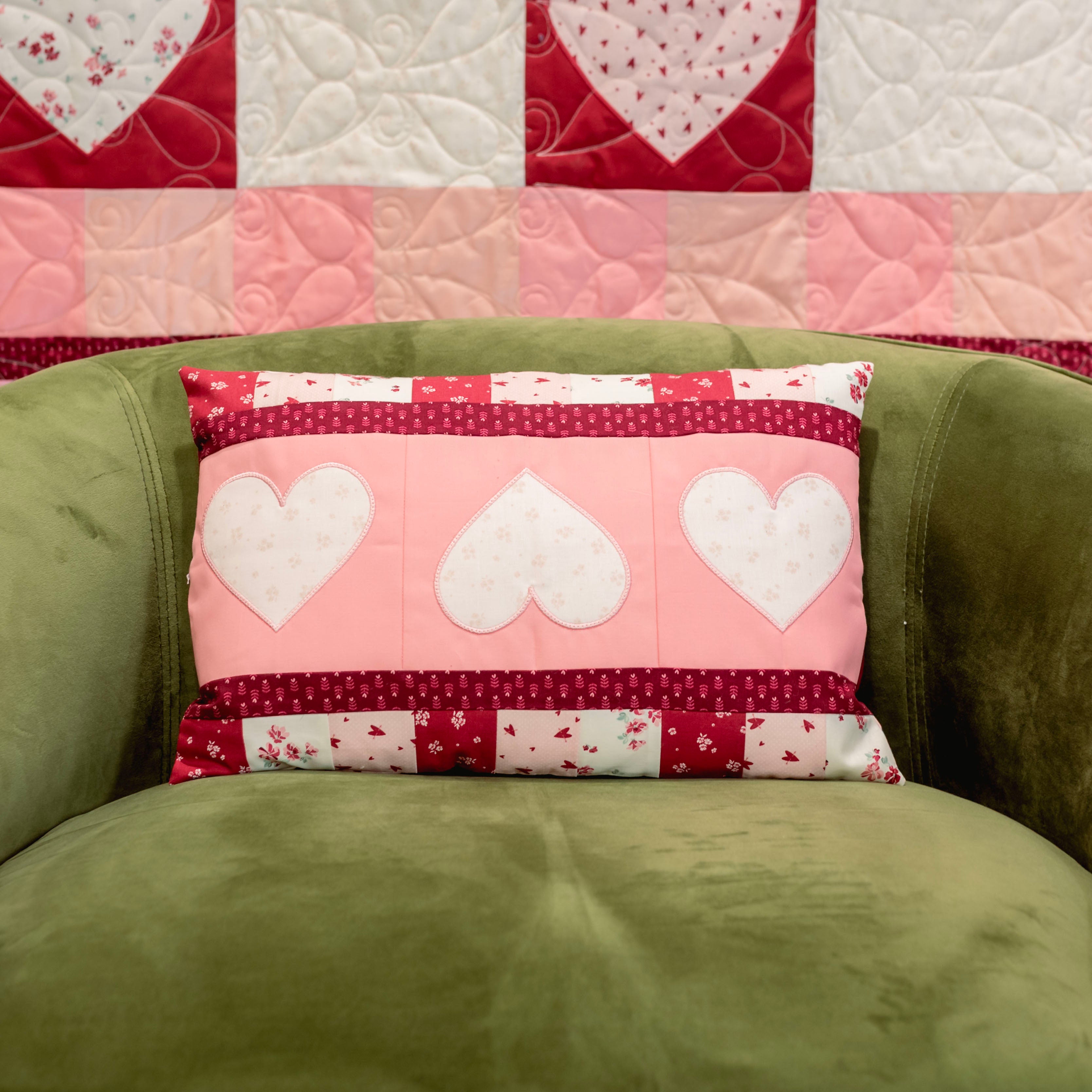 Patchwork pillow with heart designs on a green couch against a quilted wall.