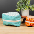 Two small zippered fabric pouches displayed on a wooden tabletop, with a potted plant in the background. One pouch is aqua blue with white polka dots and a white patterned top, and the other is orange with a small floral print and a beige patterned top.
