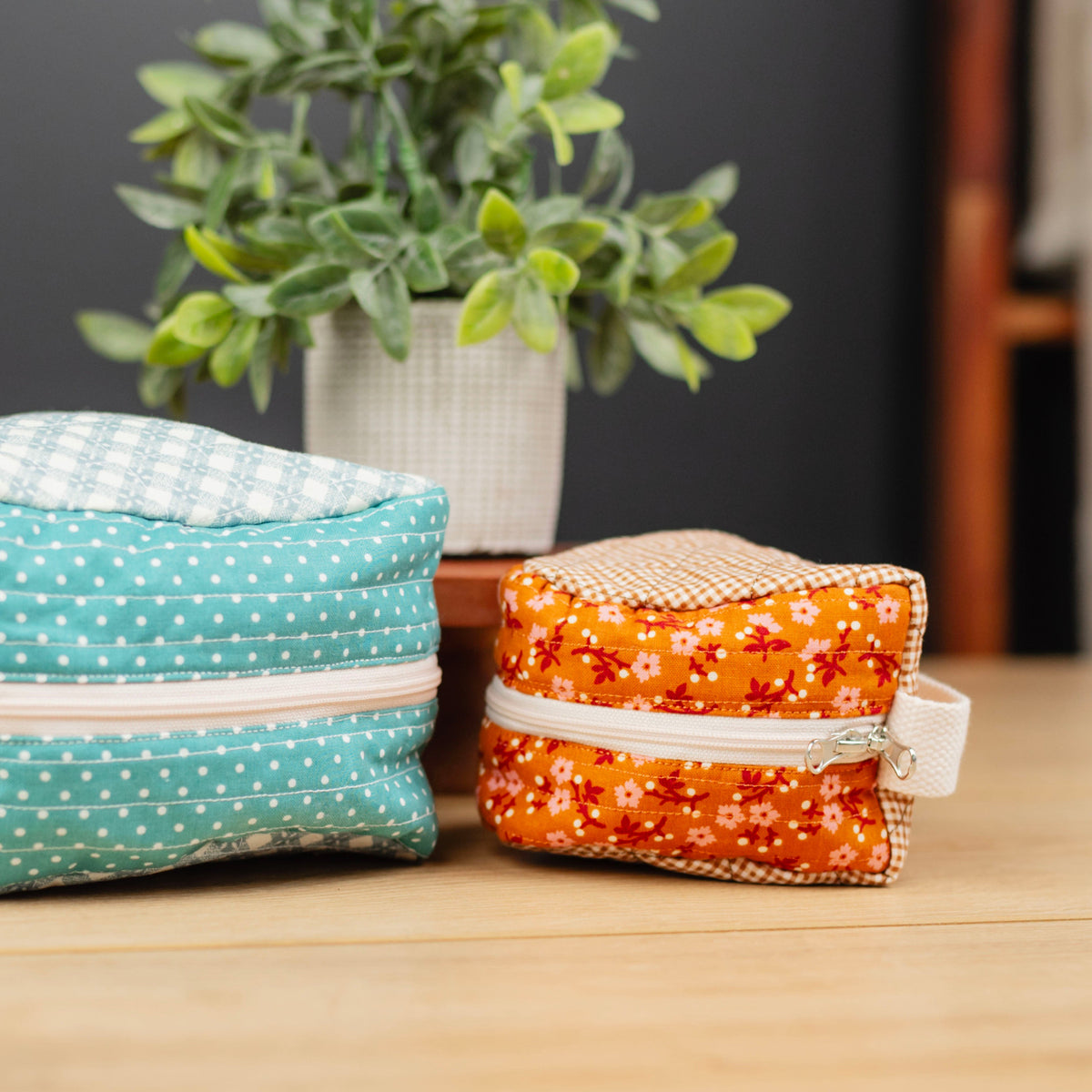 Two small zippered fabric pouches or cosmetic cases on a wooden tabletop, with a potted plant and a dark wall in the background. The left pouch is aqua blue with white polka dots and a white gingham top panel.
