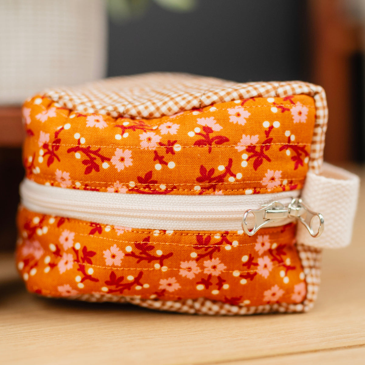 A close-up of a small zippered fabric pouch with an orange floral print, cream gingham trim, and a white zipper pull, sitting on a wooden surface next to another teal polka-dot pouch.