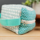 A small fabric zippered pouch with a teal and white gingham pattern on one side, a teal polka-dot pattern on the zipper flap, and a teal woven handle, resting on a wooden surface.