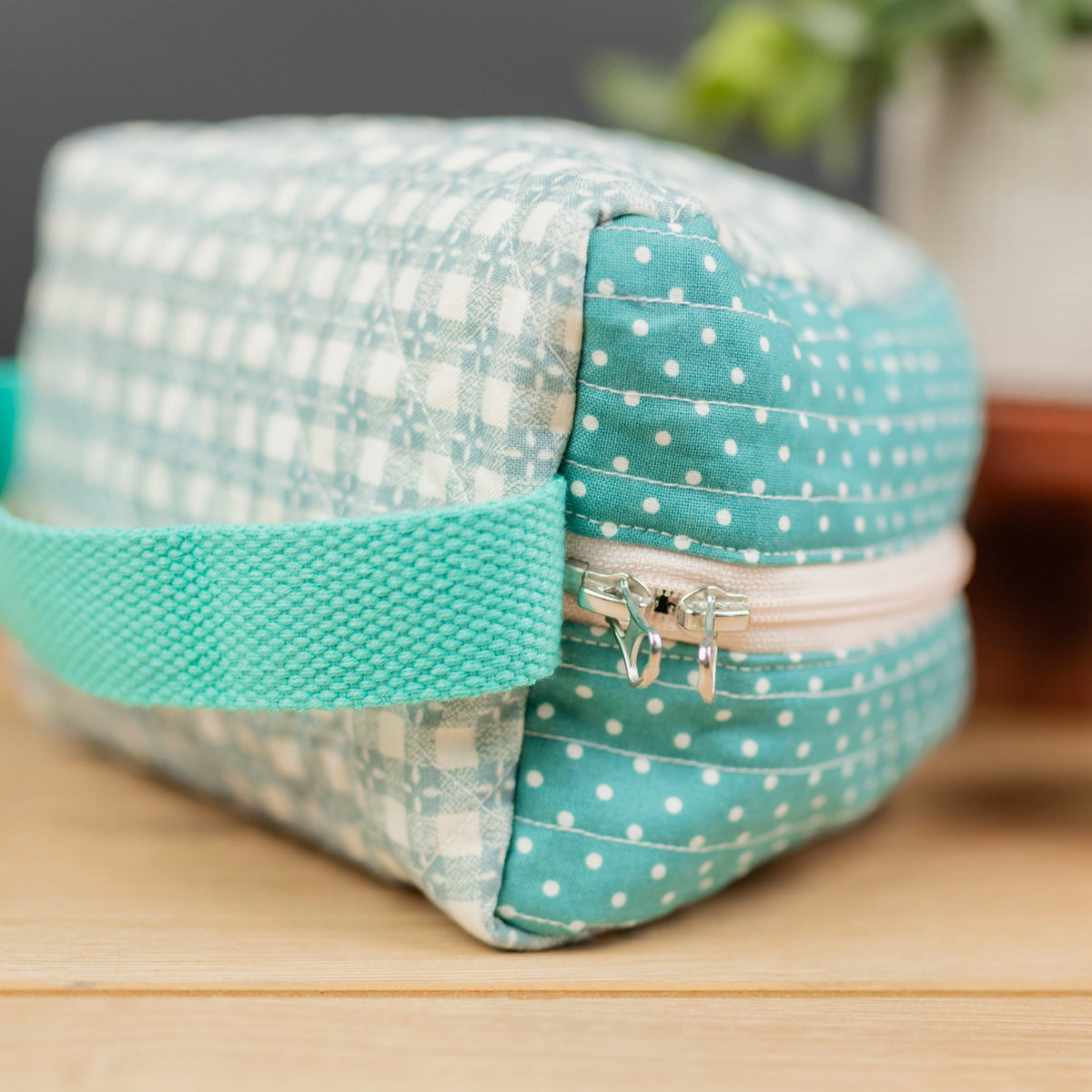A small fabric zippered pouch with a teal and white gingham pattern on one side, a teal polka-dot pattern on the zipper flap, and a teal woven handle, resting on a wooden surface.