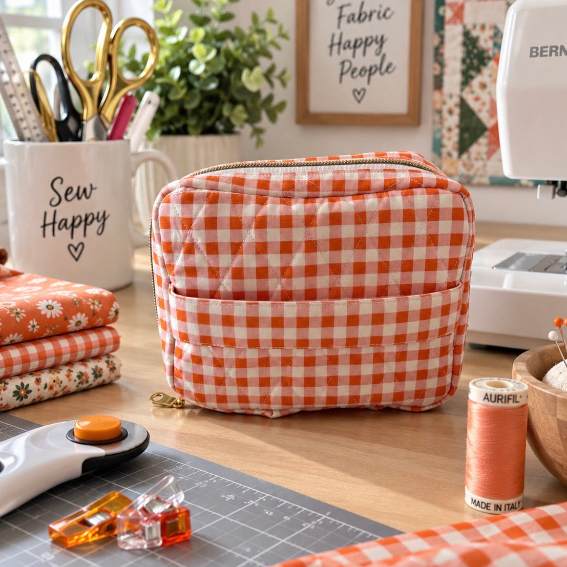 An orange and white gingham quilted zippered pouch with a front pocket, styled on a wooden table in a sewing room alongside a sewing machine, scissors, fabric, and a sewing mug.