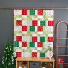 A Christmas-themed patchwork quilt hanging on a gold rod against a dark gray wall, styled with a small decorated Christmas tree, a wooden ladder, and a wooden storage bench.