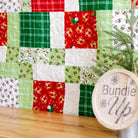 A Christmas-themed quilted fabric bundle display featuring red, green, and white patterned patches, styled with a small pine tree and a round wooden sign that reads 'Bundle Up' on a metal stand.
