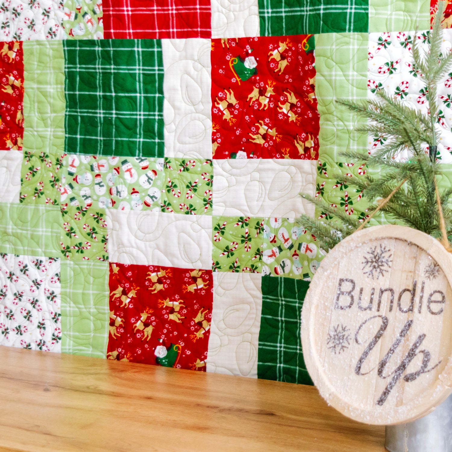 A Christmas-themed quilted fabric bundle display featuring red, green, and white patterned patches, styled with a small pine tree and a round wooden sign that reads 'Bundle Up' on a metal stand.
