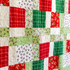 A close-up of a handmade quilt made of patchwork fabric squares in Christmas colors, featuring red, green, and white patterns including plaid, holly, pine trees, and holiday motifs.