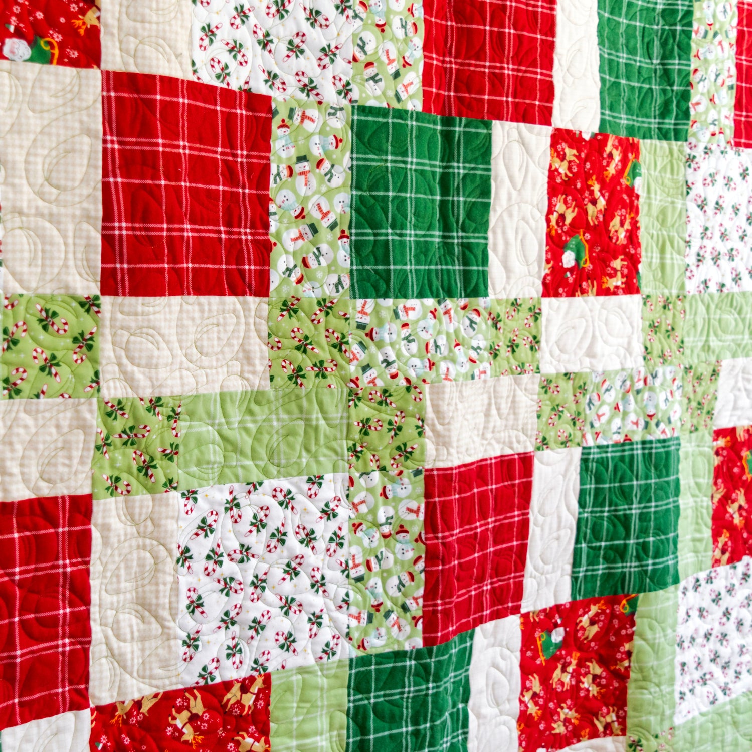 A close-up of a handmade quilt made of patchwork fabric squares in Christmas colors, featuring red, green, and white patterns including plaid, holly, pine trees, and holiday motifs.