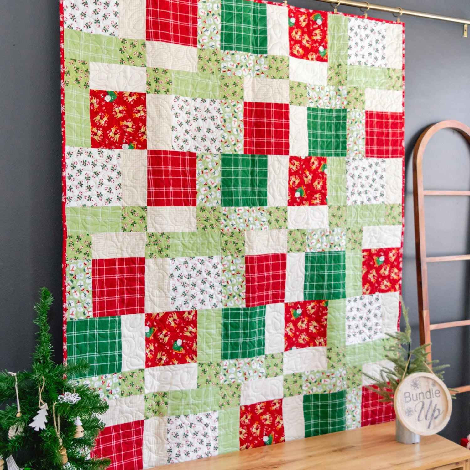 A handmade quilted patchwork blanket with a Christmas theme, featuring red, green, and white fabrics, hanging on a dark gray wall next to a wooden ladder and a small decorated tree.