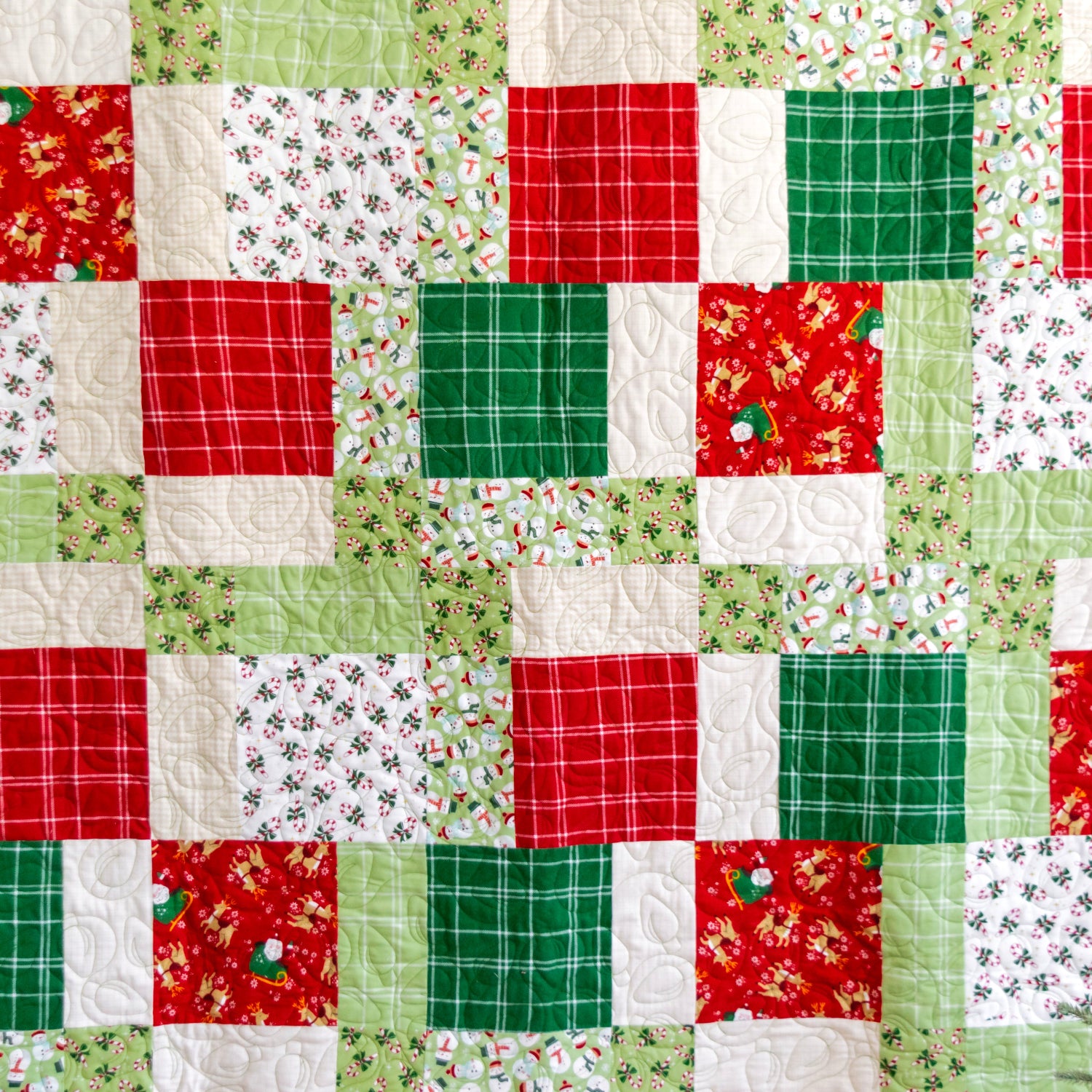 A handmade quilt with a festive holiday theme, featuring a patchwork design of red, green, and white fabric squares with various patterns including plaid, floral, and snow globes.