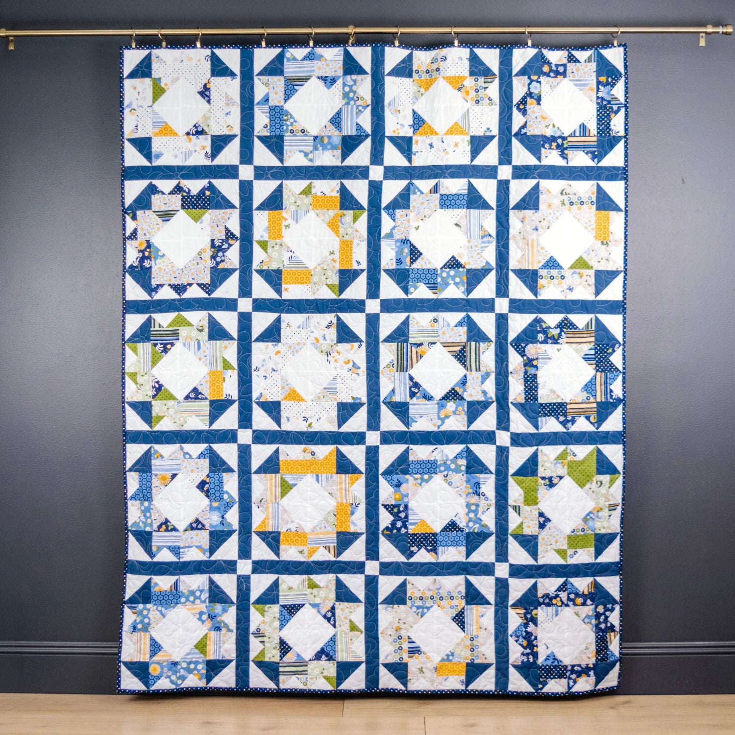 A handmade patchwork quilt hanging from a brass rod against a dark charcoal gray wall, featuring a grid of blue, white, yellow, and green geometric blocks.