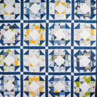 A handmade quilt featuring a grid pattern of blue, white, and yellow geometric blocks, photographed flat against a dark gray background.