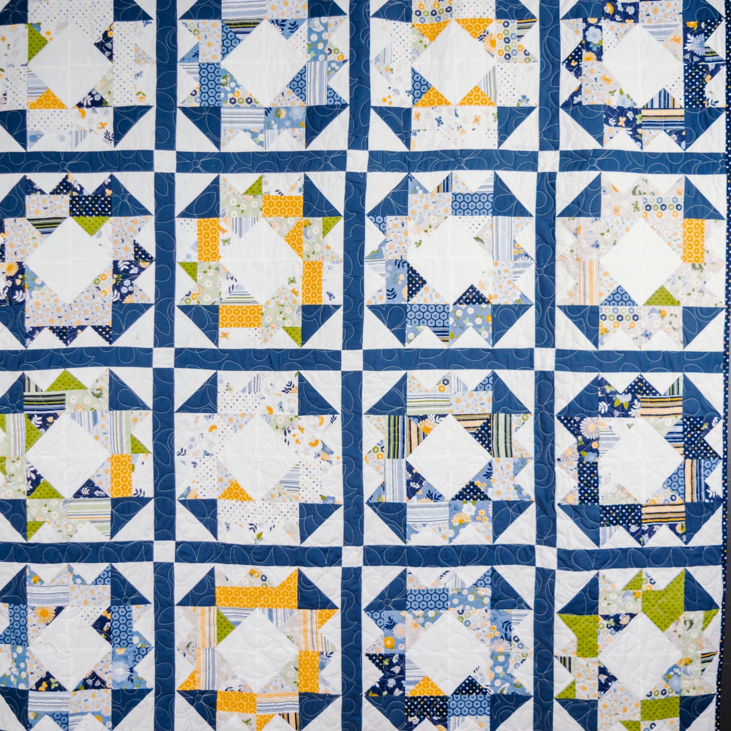 A handmade quilt featuring a grid pattern of blue, white, and yellow geometric blocks, photographed flat against a dark gray background.