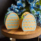 Two decorative Easter egg boxes with zigzag patterns on a wooden surface with flowers in the background.