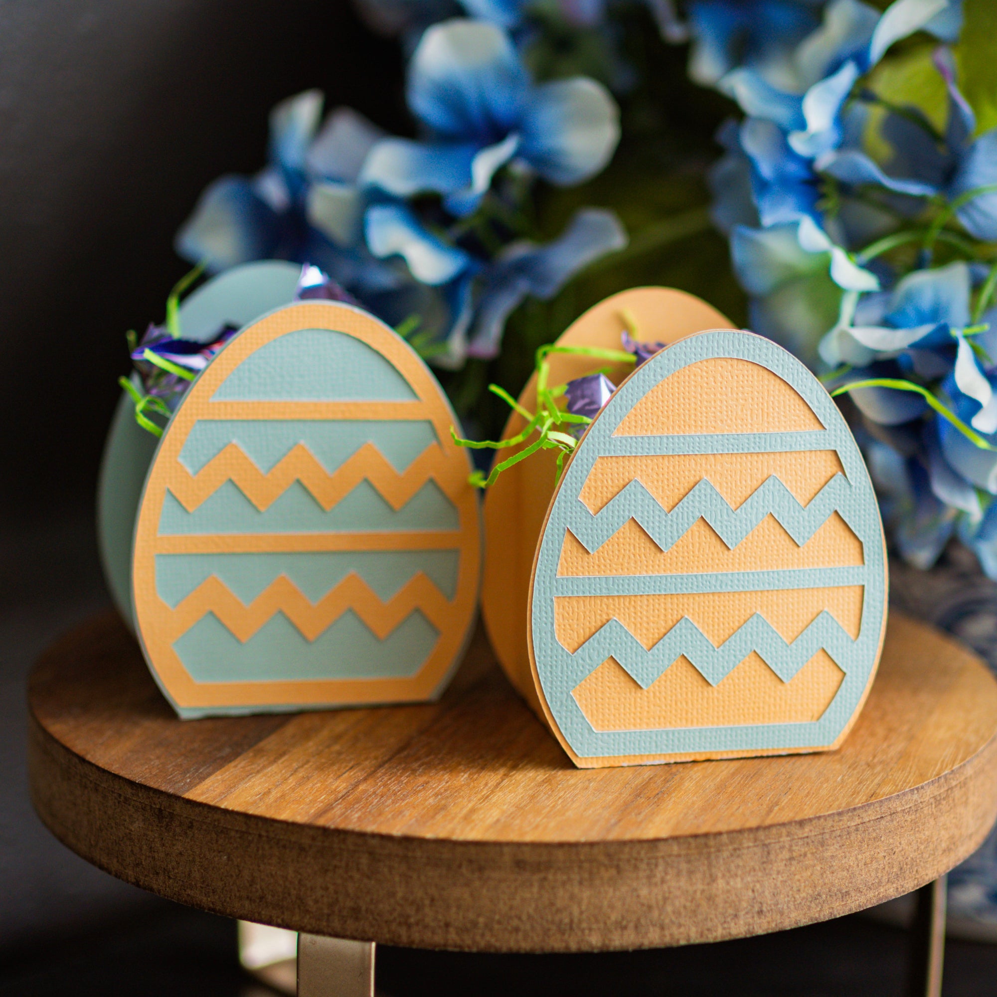 Two decorative Easter egg boxes with zigzag patterns on a wooden surface with flowers in the background.