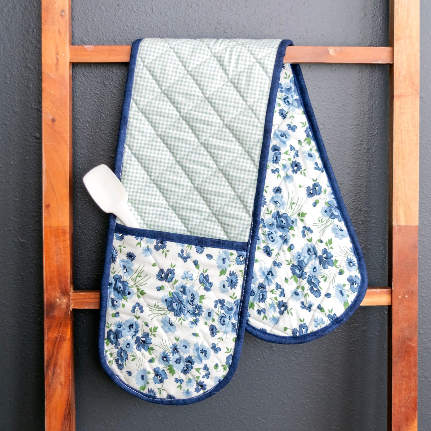 Pair of floral oven mitts hanging on a wooden rack against a gray wall.