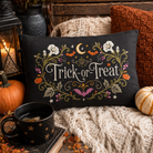 A rectangular black decorative pillow with embroidered 'Trick-or-Treat' text and autumn motifs, styled on a knit blanket with pumpkins, a mug, and a lantern in a cozy fall setting.