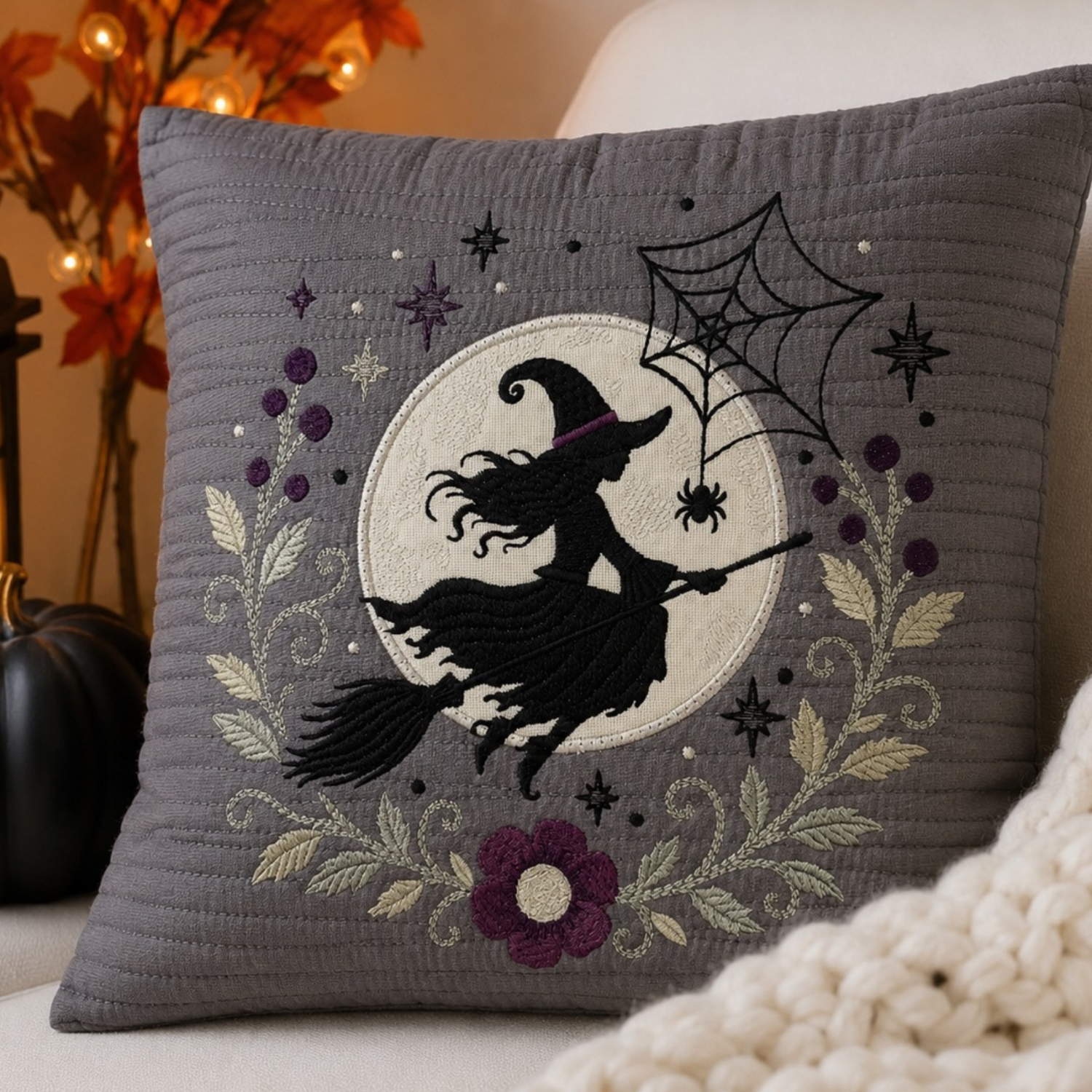 A square gray throw pillow with a Halloween-themed embroidered design of a witch riding a broomstick in front of a moon, surrounded by spiderwebs, stars, and purple flowers. The pillow is styled on a light-colored couch next to a black pumpkin and a chunky knit blanket.