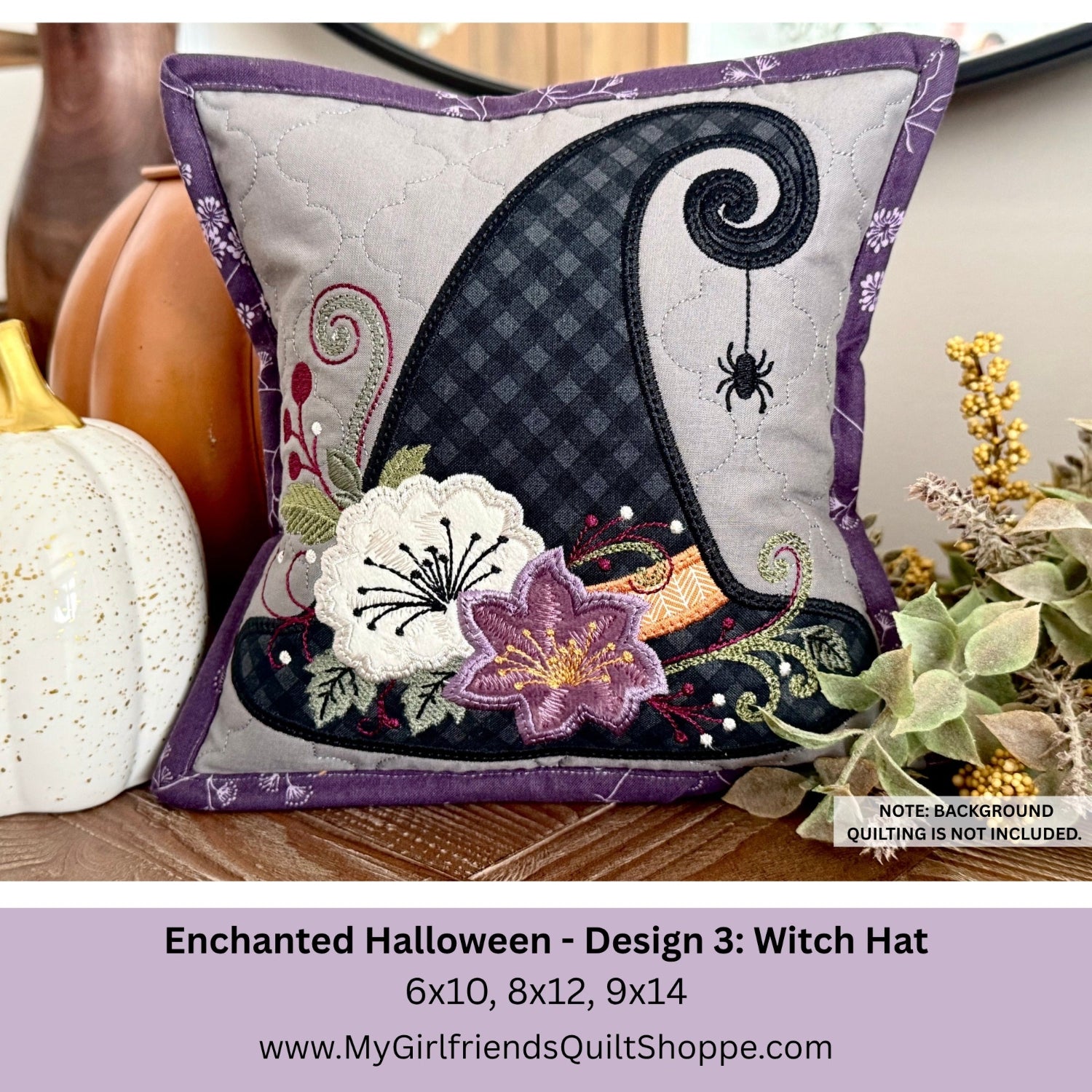 A decorative square quilted pillow shown in a styled home setting with pumpkins and dried flowers. The pillow features an applique witch hat design in black and gray with purple trim, embroidered flowers, and a spider motif.