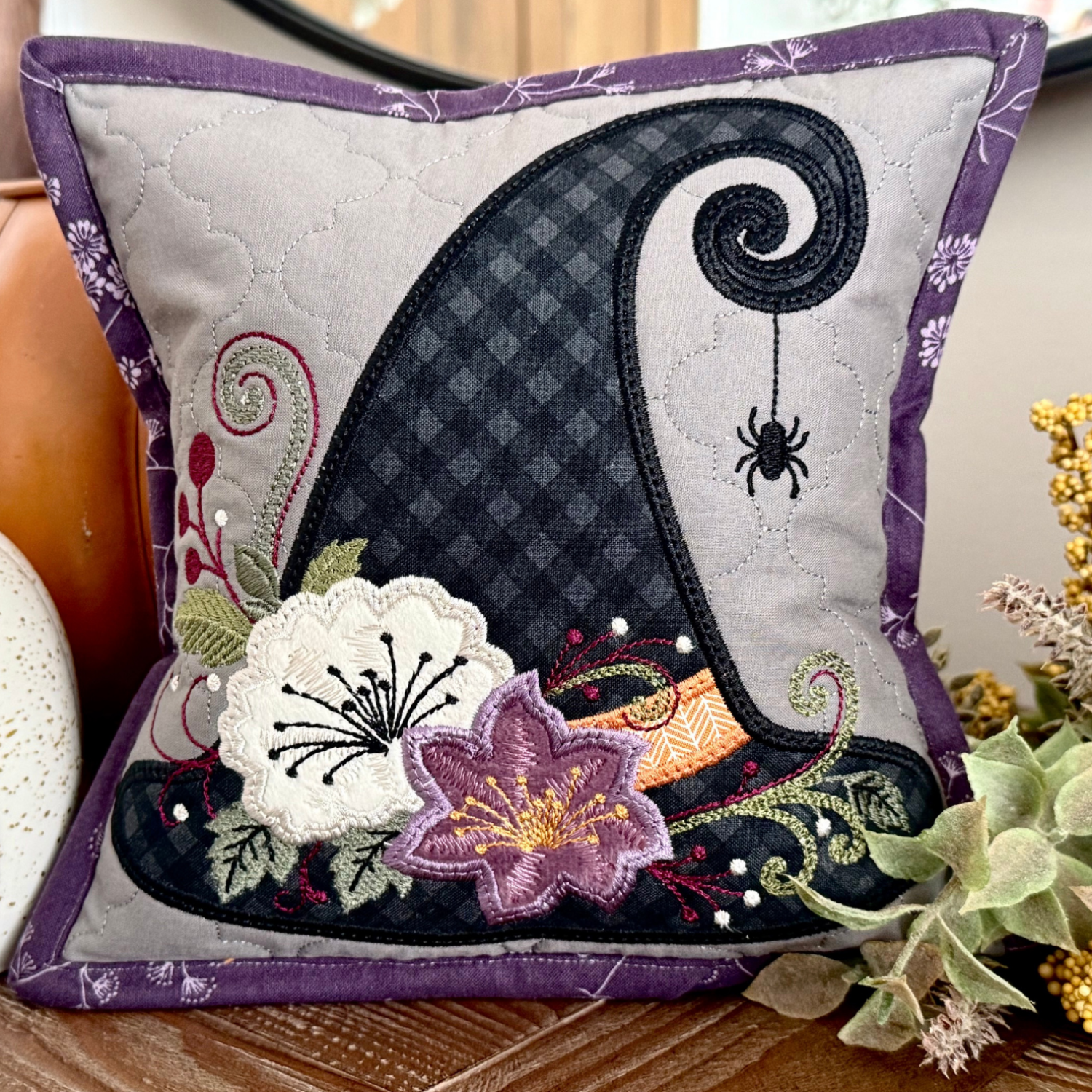 A decorative square throw pillow with a Halloween witch hat design, styled on a wooden surface with pumpkins and dried foliage.