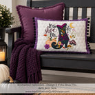 A decorative square throw pillow displayed on a dark wooden bench, styled with a matching purple pillow and a chunky knit purple throw blanket. The front pillow features a white center with a colorful embroidered design of an enchanted shoe, pumpkins, a spiderweb, and the text 'If the shoe fits.