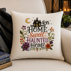 A square cream throw pillow with a colorful embroidered 'Home Sweet Haunted Home' design, styled on a beige armchair next to a black patterned throw blanket and books.