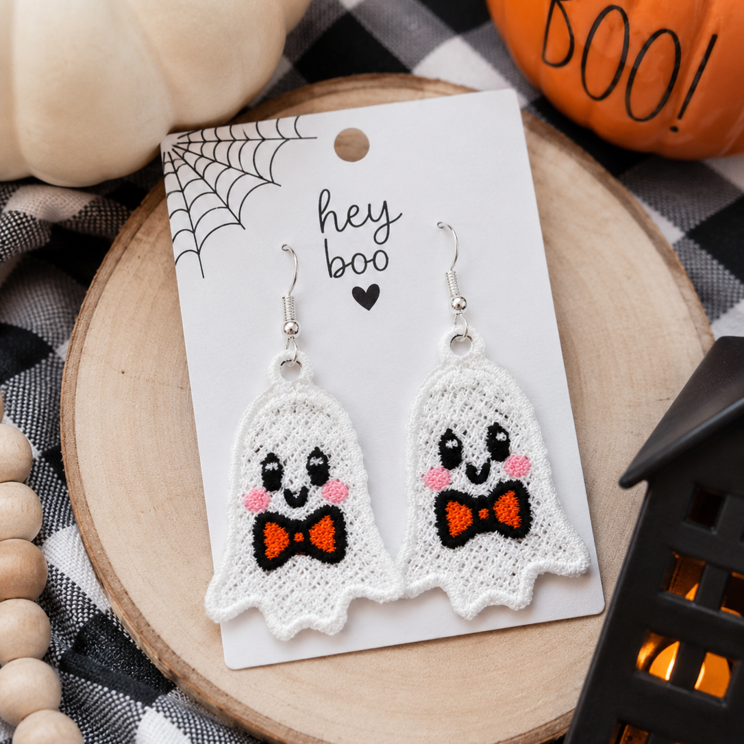 A pair of white ghost-shaped dangle earrings with embroidered smiling faces, pink cheeks, and orange bow ties, displayed on a white card that reads 'hey boo' with a small heart. The card is placed on a round wooden slice over a black-and-white checkered fabric, with decorative pumpkins, beads, and a black lantern in the background.