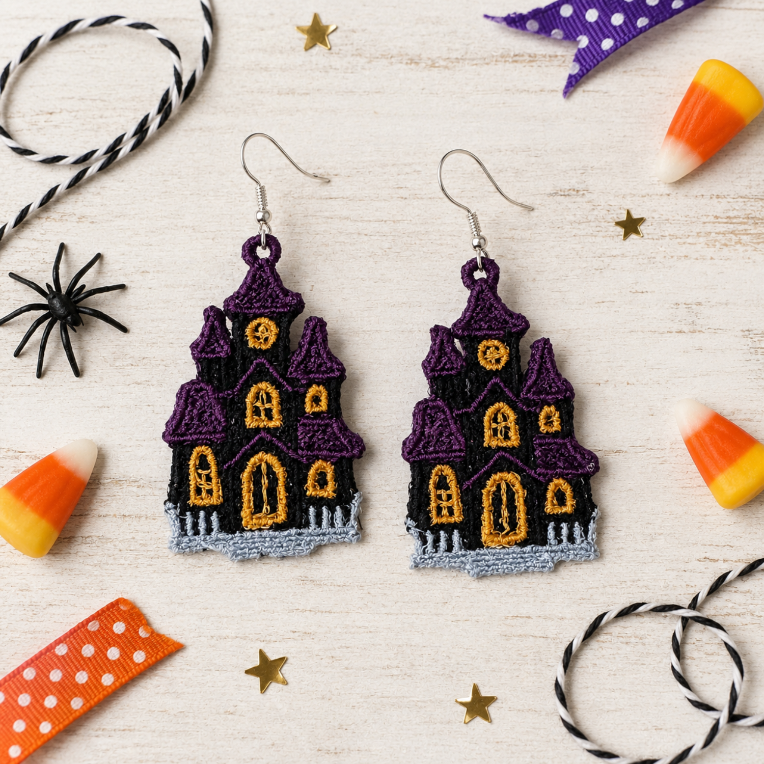 A pair of embroidered Halloween earrings shaped like haunted houses, featuring purple roofs, a black body, and glowing orange windows, displayed on a light wood background with candy corn, spider decorations, and ribbon.