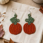 A pair of handmade embroidered pumpkin earrings with green stems and silver fishhook wires, displayed on a textured cream linen cloth with autumn props like a small white pumpkin, cinnamon sticks, and leaves.