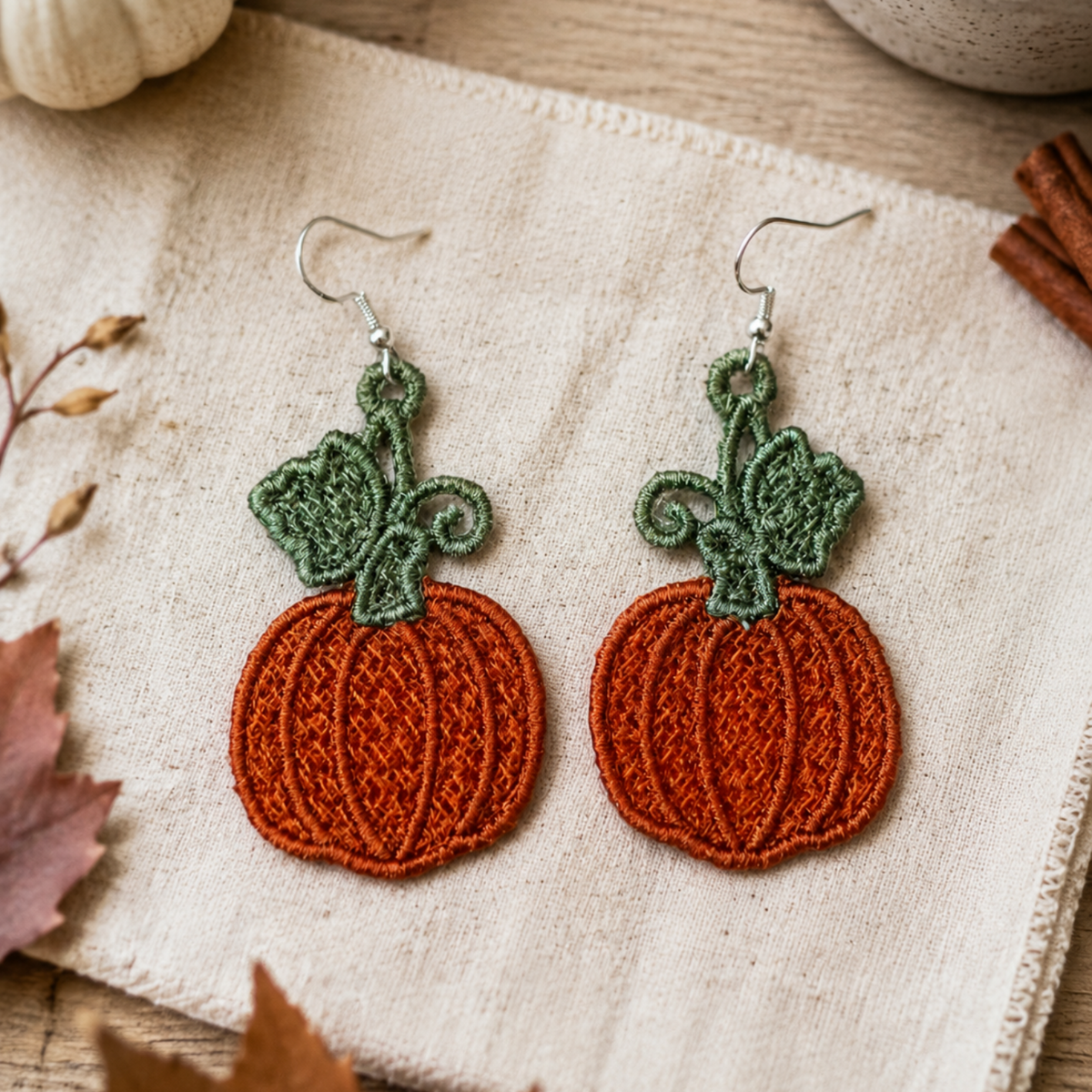 A pair of handmade embroidered pumpkin earrings with green stems and silver fishhook wires, displayed on a textured cream linen cloth with autumn props like a small white pumpkin, cinnamon sticks, and leaves.