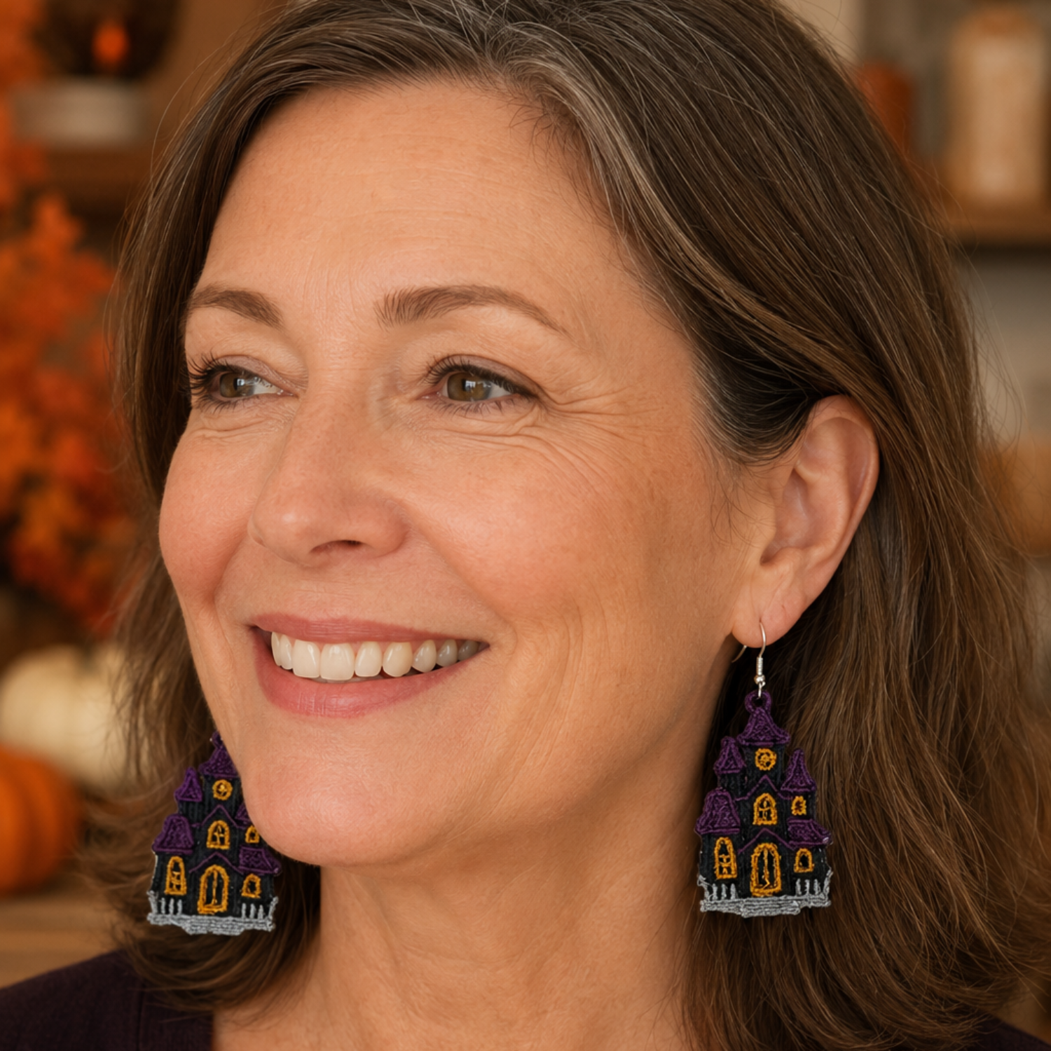 A smiling woman with shoulder-length brown hair wearing large, colorful Halloween-themed dangle earrings shaped like haunted houses. The earrings feature purple roofs, yellow windows, and gray details, shown against a softly blurred warm indoor background.