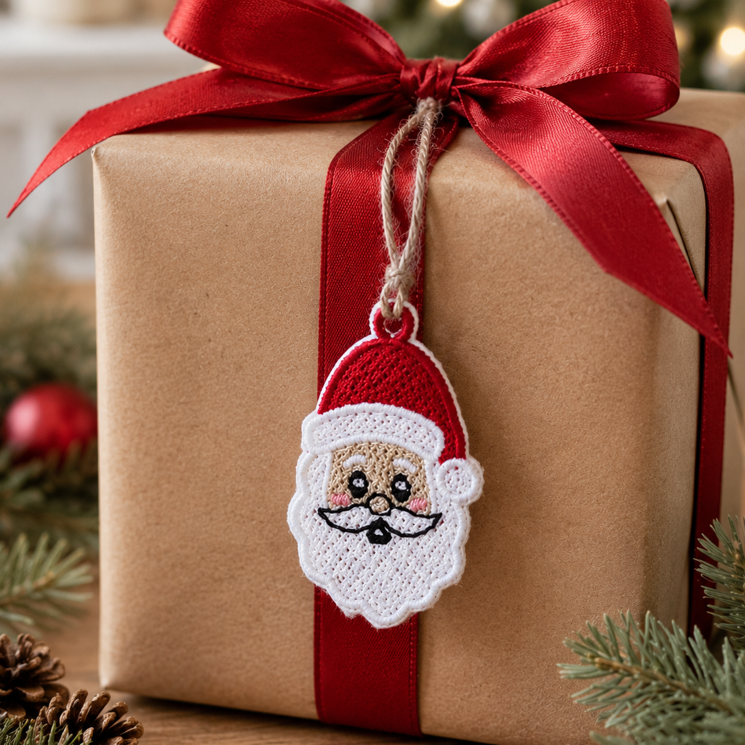 A wrapped gift box in brown paper with a large red ribbon and bow, featuring a hanging embroidered Santa face ornament. The scene is styled with pine branches, a pinecone, and a softly blurred Christmas tree in the background.