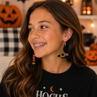 A smiling woman with long brown hair and braces wearing colorful dangling earrings and a black graphic sweatshirt that reads 'Hocus Pocus' in white, set against a cozy Halloween-themed background with pumpkins and warm lights.