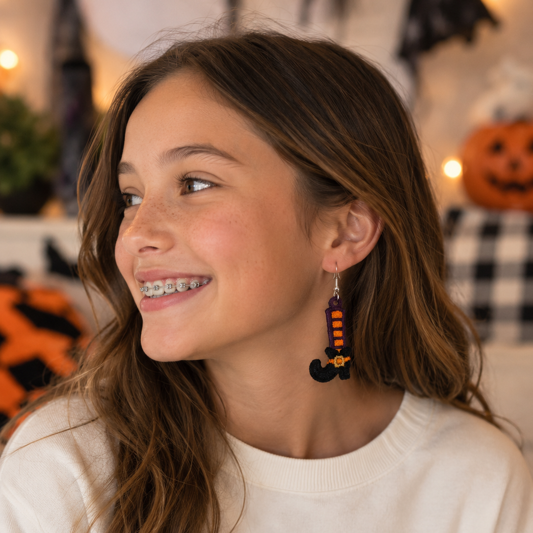 A smiling young woman with long brown hair is shown in profile wearing a pair of dangling witch hat earrings. She is wearing a cream sweatshirt with a 'just a girl who loves' graphic, set against a cozy, Halloween-decorated background with warm string lights and pumpkins.