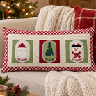 A rectangular Christmas throw pillow with a red and white gingham border and three central holiday panels showing Santa, a Christmas tree, and a snowman, styled on a beige couch with a knit blanket in a cozy living room setting.