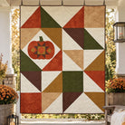 A large quilted wall hanging or banner with an autumn geometric pattern and a pumpkin motif, displayed on a covered porch with outdoor foliage visible in the background.