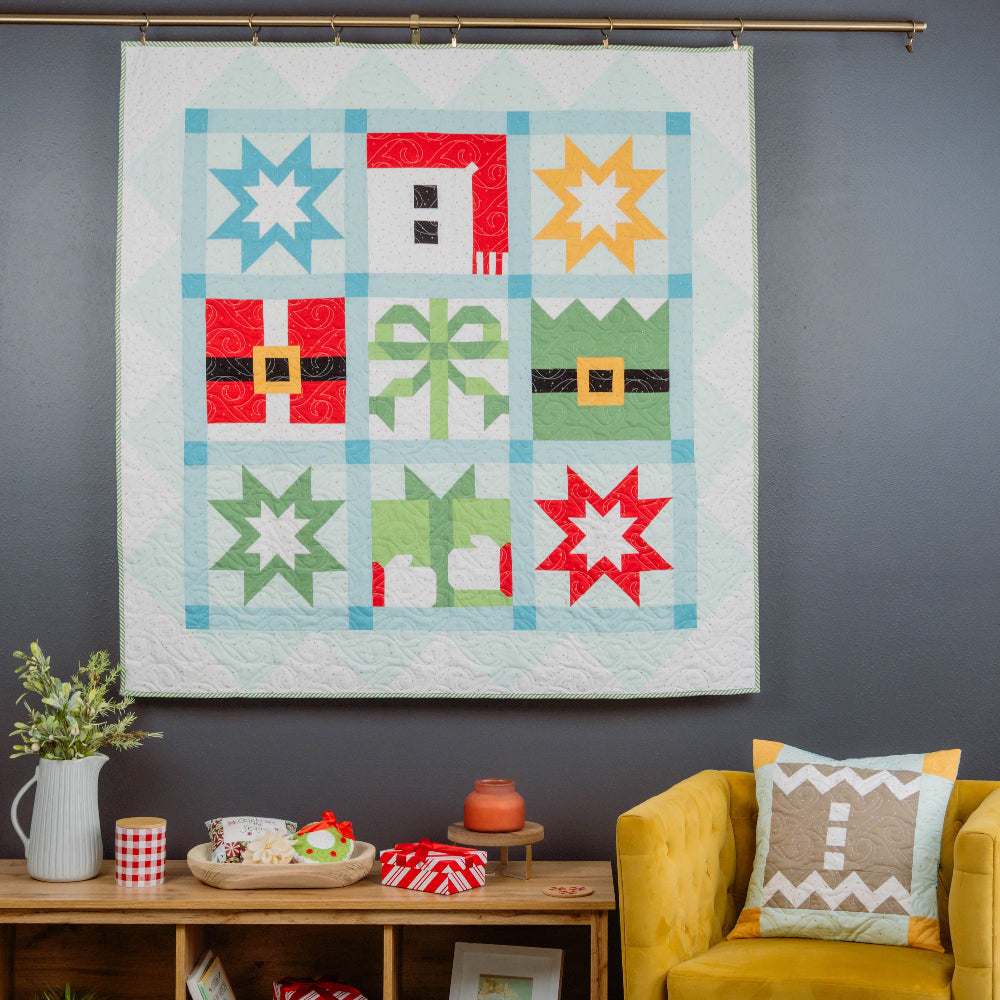 Colorful quilt with geometric patterns hanging on a gray wall above a yellow armchair.