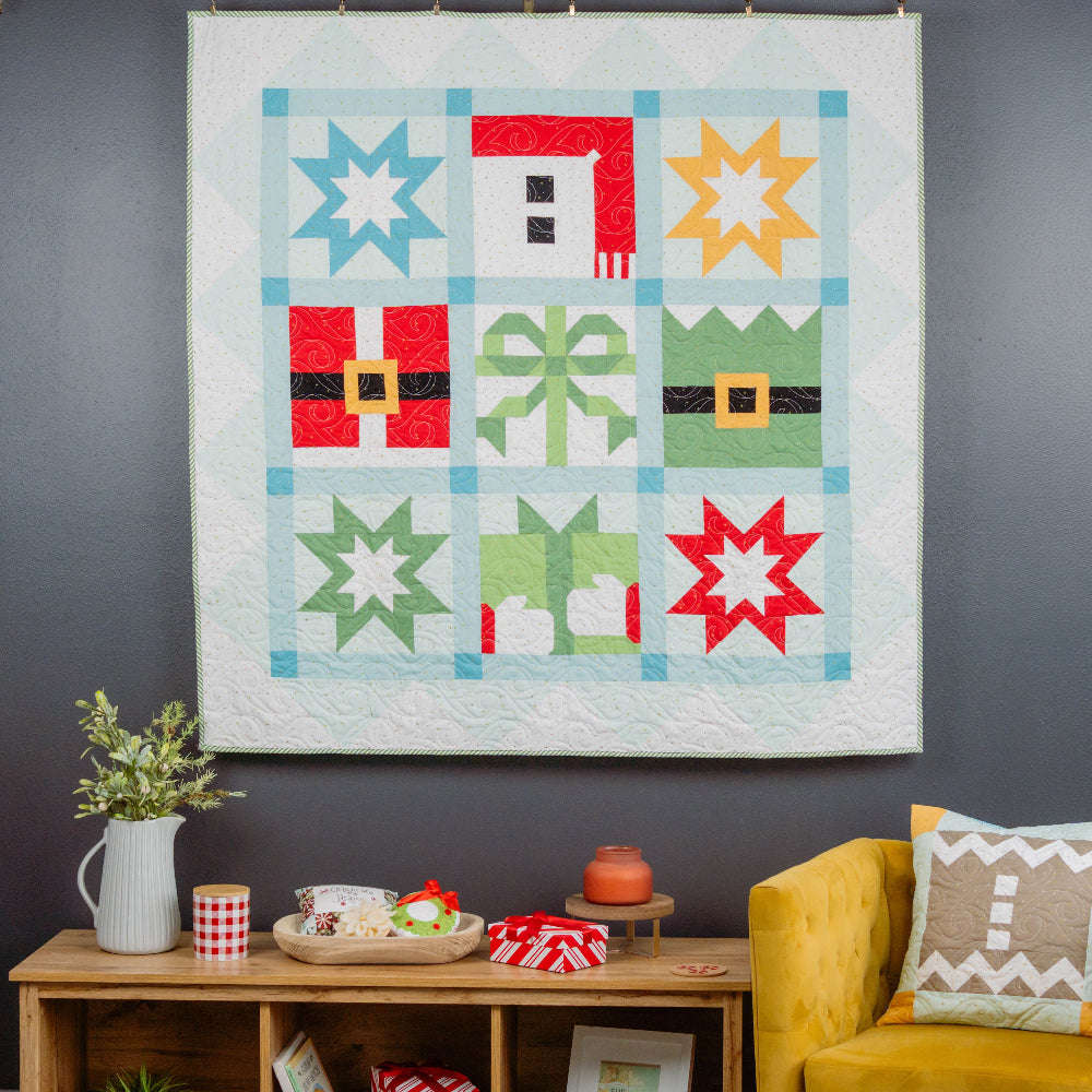 Colorful quilt hanging on a dark wall above a wooden shelf with decorative items and a yellow chair.