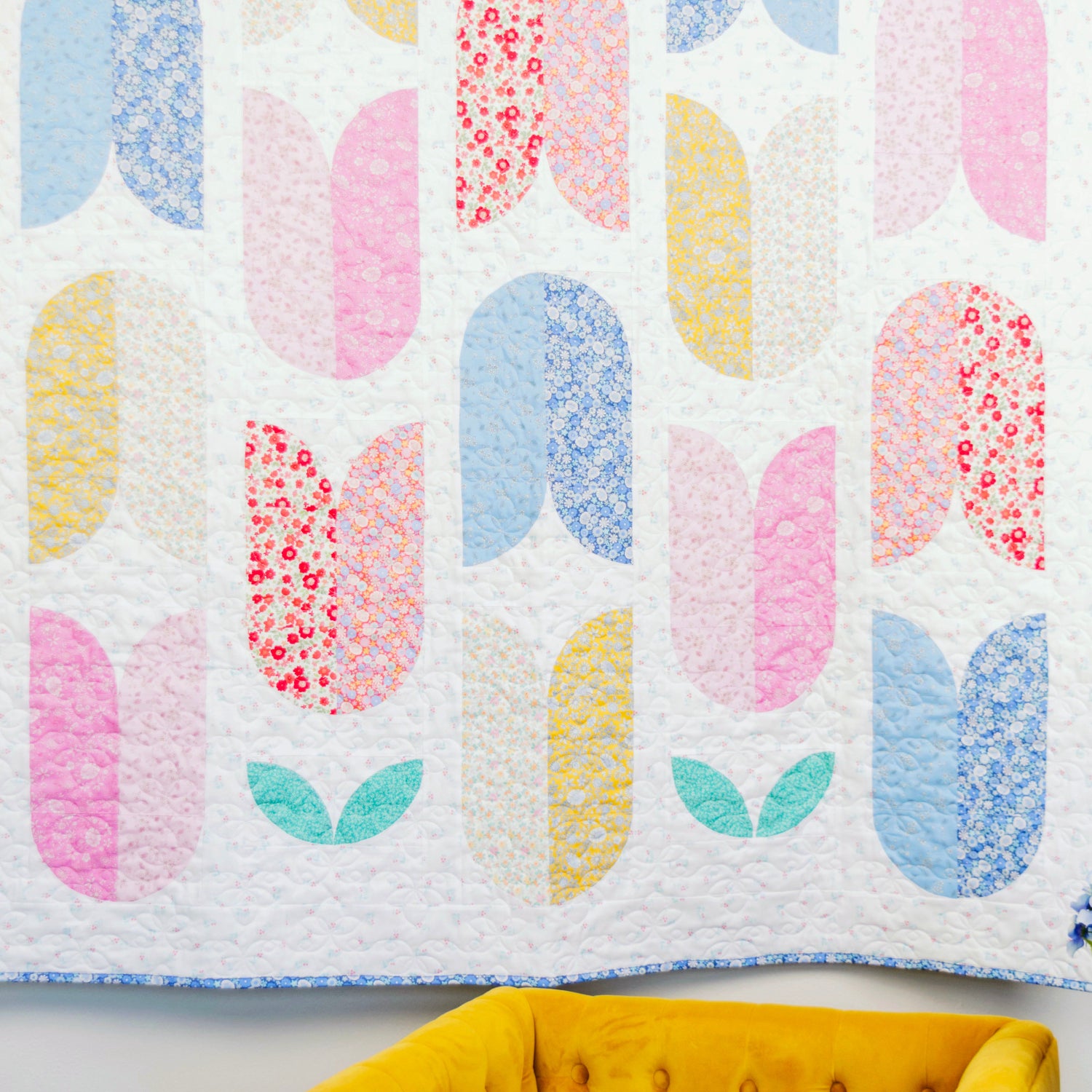 Colorful quilt with abstract pattern on a white wall with a yellow chair and blue flowers.