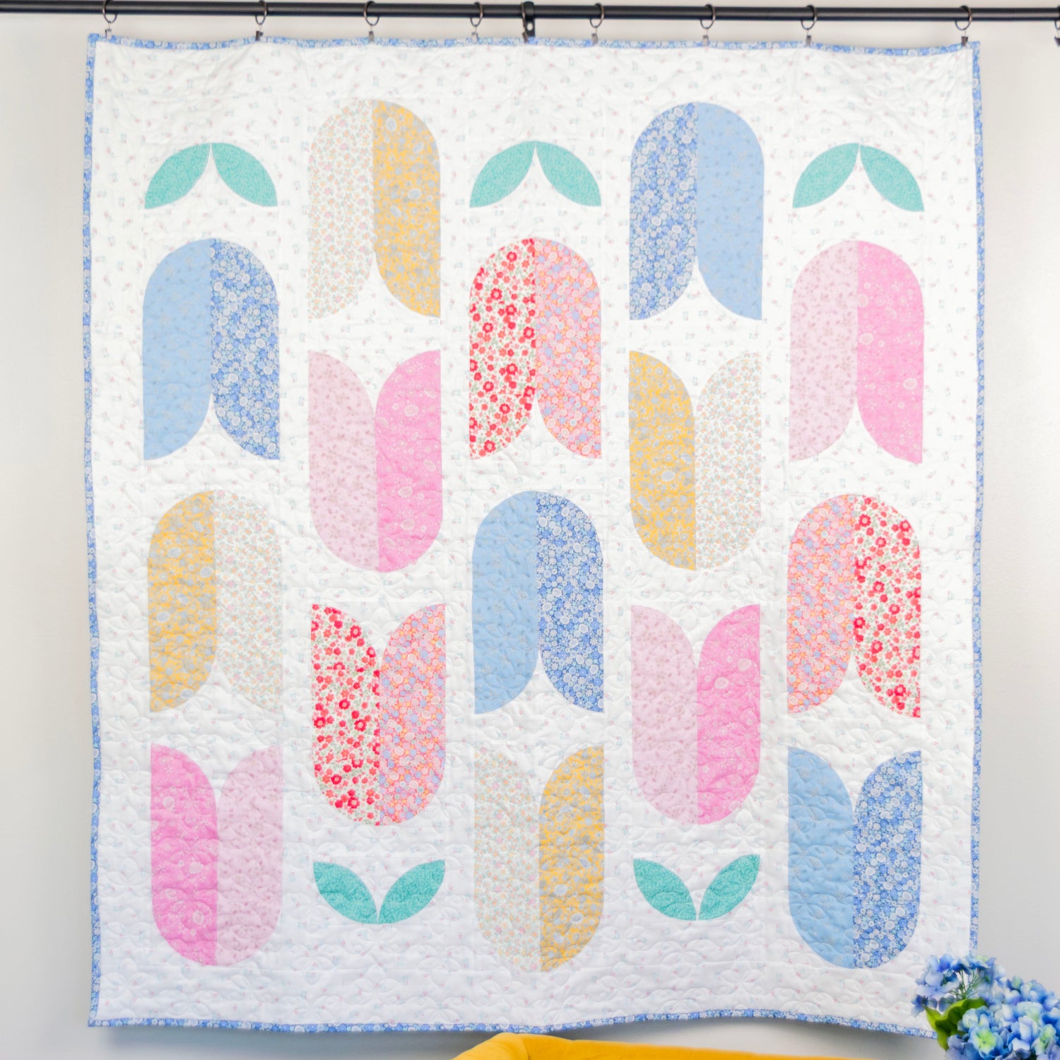 Colorful quilt with floral patterns hanging on a wall above a yellow chair.