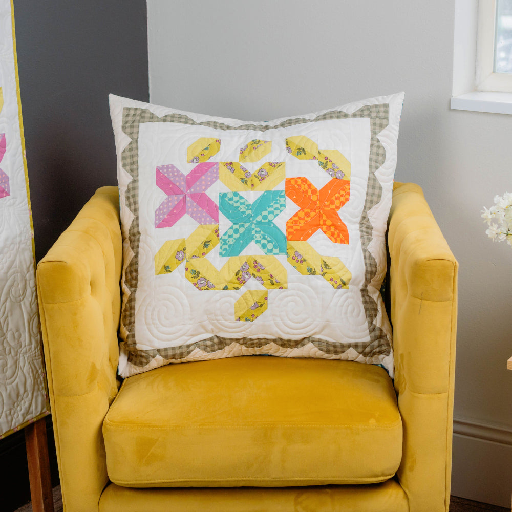 Yellow armchair with a decorative pillow featuring colorful geometric patterns, next to a quilt and a small table with flowers.