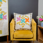 Yellow armchair with a quilted pillow featuring colorful floral patterns, next to a quilt on a stand against a gray wall.