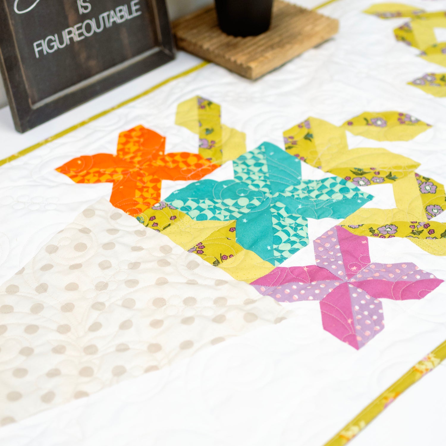 Colorful quilt block on a white surface with a motivational sign in the background.