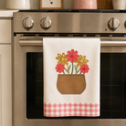 A white kitchen towel with a floral pot design and pink gingham hem hanging on a stainless steel oven handle in a decorated kitchen.