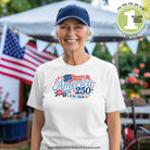 Woman wearing a white t-shirt with 'America 250' design, standing outdoors with flags in the background.